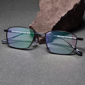 May include: Black rectangular eyeglasses with a thin frame and gradient lenses. The glasses are resting on a grey surface with a rock in the background. The text "TITANIUM 145 C4" is visible on the frame.