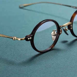 May include: Close-up of round eyeglasses with a dark brown tortoiseshell frame and gold-coloured metal accents. The lenses are clear, and the glasses have a classic, vintage aesthetic. The frame is a dark brown colour with a tortoiseshell pattern.