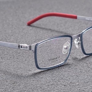 May include: Close-up of eyeglasses with a rectangular frame. The frame is silver with a blue outline and red temple tips. The text "TITANIUM IP" and measurements are visible on the frame.