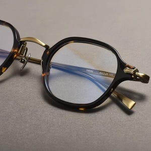 May include: Close-up of eyeglasses with a dark brown tortoiseshell frame and gold-colored accents. The lenses are clear, and the frame has a square shape. The text "HAVAG 43☐24-145" is visible on the frame.