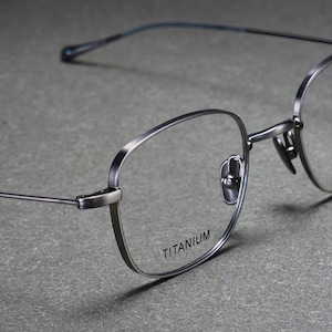 Puede incluir: Primer plano de gafas de color plateado con montura rectangular redondeada. El texto "TITANIUM" está impreso en la parte inferior de la montura. Las gafas tienen patillas finas y delicadas y almohadillas nasales.