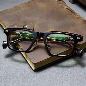 Puede incluir: Gafas rectangulares negras y carey. Las gafas, hechas de titanio, descansan sobre un libro encuadernado en cuero marrón. Las lentes tienen un ligero tinte verde.
