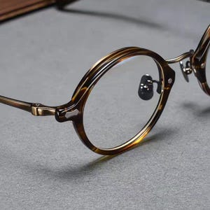Puede incluir: Gafas redondas con montura de patrón de carey marrón y patillas de metal en tono bronce. Las gafas incluyen lentes transparentes, almohadillas nasales ajustables y detalles texturizados en el puente y las uniones de las patillas.