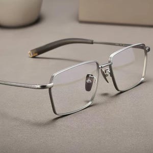 Könnte beinhalten: Silberfarbene Brille mit rechteckiger Gläserform. Die Brille hat ein strukturiertes Muster auf dem Rahmen und den Bügeln, mit einem dunkelgrauen Bügel und Nasenpads. Die Gläser sind klar, und das Design ist modern und anspruchsvoll.
