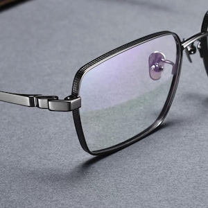 Könnte beinhalten: Eine Brille mit einem dunkelgrauen Metallrahmen, der am äußeren Rand ein texturiertes Kreuzschraffurmuster aufweist. Die Brille hat klare Gläser, dünne Bügel und integrierte Nasenpads, was ein strukturiertes und geometrisches Design ergibt.