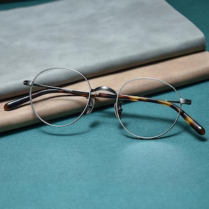 Puede incluir: Gafas redondas con montura de metal plateado y patillas carey. Las gafas descansan sobre una superficie turquesa con un fondo gris y beige. La palabra "TITANIUM" está impresa en la montura.