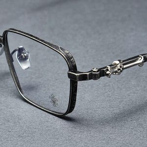 Puede incluir: Primer plano de gafas con montura metálica oscura. Las gafas tienen forma cuadrada con adornos decorativos en las patillas y bisagras. Las lentes son transparentes y el diseño general tiene una estética vintage o gótica.