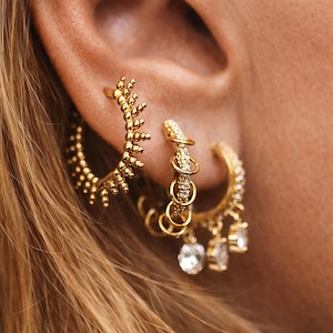 Op de afbeelding: Close-up van gouden oorbellen. Eén oorbel is een halve hoepel met een getextureerd, takachtig ontwerp. De andere is een hoepel met bungelende heldere edelstenen. De oorbellen worden gedragen op het oor van een persoon, met blond haar zichtbaar.