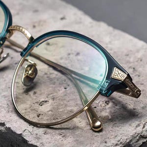 May include: Close-up of eyeglasses with a teal and gold frame. The lenses are clear, and the frame has intricate gold detailing on the temples and bridge. The glasses rest on a textured, gray surface.
