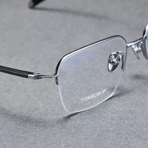 May include: Close-up of rimless eyeglasses with a silver frame and black temples. The lenses are clear, and the text "TITANIUM-IP" is printed on the lens. The glasses are set against a gray background.