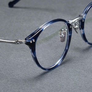 May include: Close-up of spectacles with blue and white marbled frames. The round-shaped glasses feature silver-coloured metal detailing at the hinges and nose pads. The lenses are clear.