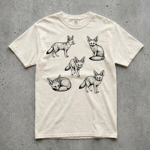 Ivory graphic t-shirt featuring a vintage engraved style illustration of multiple fennec foxes arranged in different poses. The foxes are shown sitting, walking, and curled up, with detailed linework creating a naturalist wildlife illustration and desert animal aesthetic. Laid flat on a concrete surface.