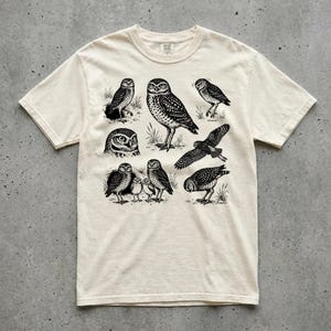 May include: A cream-colored short-sleeve t-shirt featuring a black ink-style graphic print of various burrowing owls in different poses, including standing, flying, and perched, with detailed feather patterns and small tufts of grass.