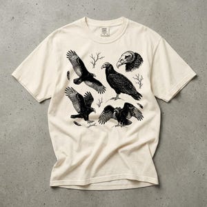 Ivory Comfort Colors t-shirt with a vintage engraved illustration of five vultures including one soaring with wings outstretched, one standing with wings half-spread, a close-up portrait showing bare head and hooked beak, one landing with wings fanned, and one spreading wings low in a dominance display, surrounded by bare winter branches and scattered feathers in an antique wildlife field guide etching style. Laid flat on a concrete surface.