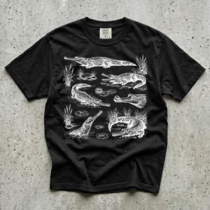 May include: Black t-shirt with a white illustration of alligators in a swamp. The design shows different alligator poses and aquatic plants. The shirt has a crew neck and short sleeves. The t-shirt is a casual wear item.