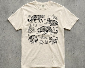May include: Cream-colored t-shirt featuring a black and white illustration of snow leopards in various poses. The design includes a walking leopard, a resting leopard, a cub, mountains, and floral elements. The shirt has a crew neck and short sleeves.
