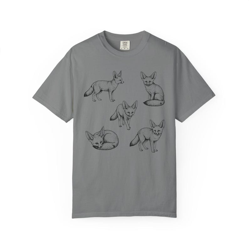 Grey graphic t-shirt featuring a vintage engraved style illustration of multiple fennec foxes arranged in different poses. The foxes are shown sitting, walking, and curled up, with detailed linework creating a naturalist wildlife illustration and desert animal aesthetic.