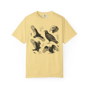 Butter yellow Comfort Colors t-shirt with a vintage engraved illustration of five vultures including one soaring with wings outstretched, one standing with wings half-spread, a close-up portrait showing bare head and hooked beak, one landing with wings fanned, and one spreading wings low in a dominance display, surrounded by bare winter branches and scattered feathers in an antique wildlife field guide etching style.