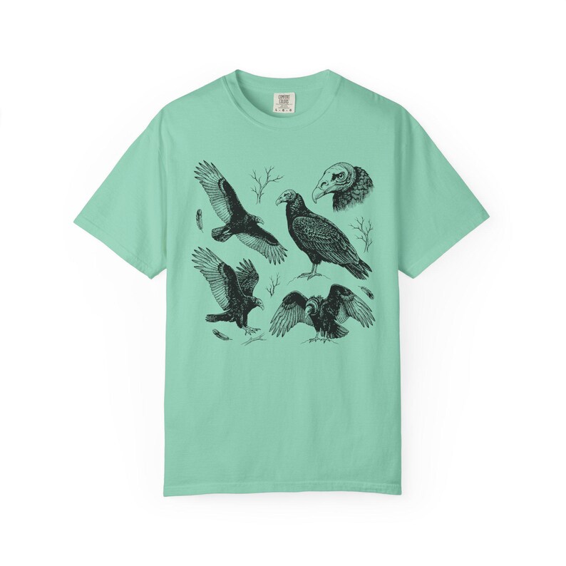 Island reef green Comfort Colors t-shirt with a vintage engraved illustration of five vultures including one soaring with wings outstretched, one standing with wings half-spread, a close-up portrait showing bare head and hooked beak, one landing with wings fanned, and one spreading wings low in a dominance display, surrounded by bare winter branches and scattered feathers in an antique wildlife field guide etching style.