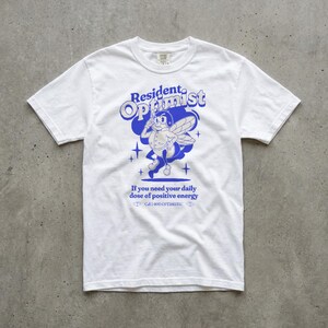 May include: White t-shirt featuring a blue cartoon bee graphic and the words "Resident Optimist." The shirt also includes the text "If you need your daily dose of positive energy" and a phone number.
