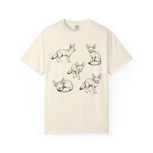 Ivory graphic t-shirt featuring a vintage engraved style illustration of multiple fennec foxes arranged in different poses. The foxes are shown sitting, walking, and curled up, with detailed linework creating a naturalist wildlife illustration and desert animal aesthetic.