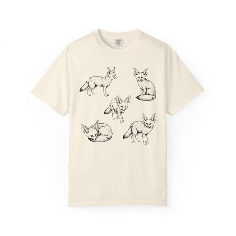 Ivory graphic t-shirt featuring a vintage engraved style illustration of multiple fennec foxes arranged in different poses. The foxes are shown sitting, walking, and curled up, with detailed linework creating a naturalist wildlife illustration and desert animal aesthetic.