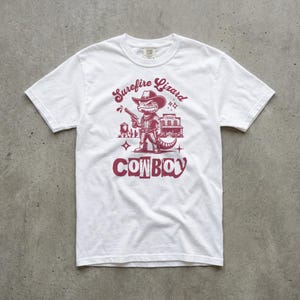 May include: White t-shirt with a maroon graphic featuring a lizard cowboy holding a gun, with the text "Surefire Lizard Cowboy". The design includes a saloon and other western elements. The shirt is made of a soft, comfortable material.