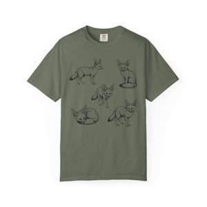 Moss green graphic t-shirt featuring a vintage engraved style illustration of multiple fennec foxes arranged in different poses. The foxes are shown sitting, walking, and curled up, with detailed linework creating a naturalist wildlife illustration and desert animal aesthetic.