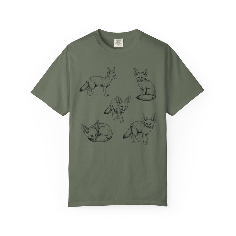 Moss green graphic t-shirt featuring a vintage engraved style illustration of multiple fennec foxes arranged in different poses. The foxes are shown sitting, walking, and curled up, with detailed linework creating a naturalist wildlife illustration and desert animal aesthetic.