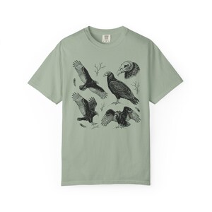 Bay green Comfort Colors t-shirt with a vintage engraved illustration of five vultures including one soaring with wings outstretched, one standing with wings half-spread, a close-up portrait showing bare head and hooked beak, one landing with wings fanned, and one spreading wings low in a dominance display, surrounded by bare winter branches and scattered feathers in an antique wildlife field guide etching style.