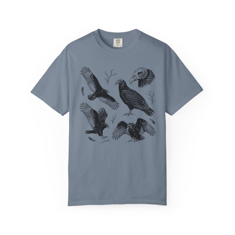 Blue jean Comfort Colors t-shirt with a vintage engraved illustration of five vultures including one soaring with wings outstretched, one standing with wings half-spread, a close-up portrait showing bare head and hooked beak, one landing with wings fanned, and one spreading wings low in a dominance display, surrounded by bare winter branches and scattered feathers in an antique wildlife field guide etching style.