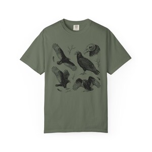 Moss green Comfort Colors t-shirt with a vintage engraved illustration of five vultures including one soaring with wings outstretched, one standing with wings half-spread, a close-up portrait showing bare head and hooked beak, one landing with wings fanned, and one spreading wings low in a dominance display, surrounded by bare winter branches and scattered feathers in an antique wildlife field guide etching style.
