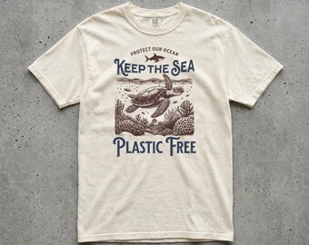 May include: Cream-colored t-shirt with a graphic design promoting ocean conservation. The design features a sea turtle, coral, and the text "Keep the Sea Plastic Free" and "Protect Our Ocean."