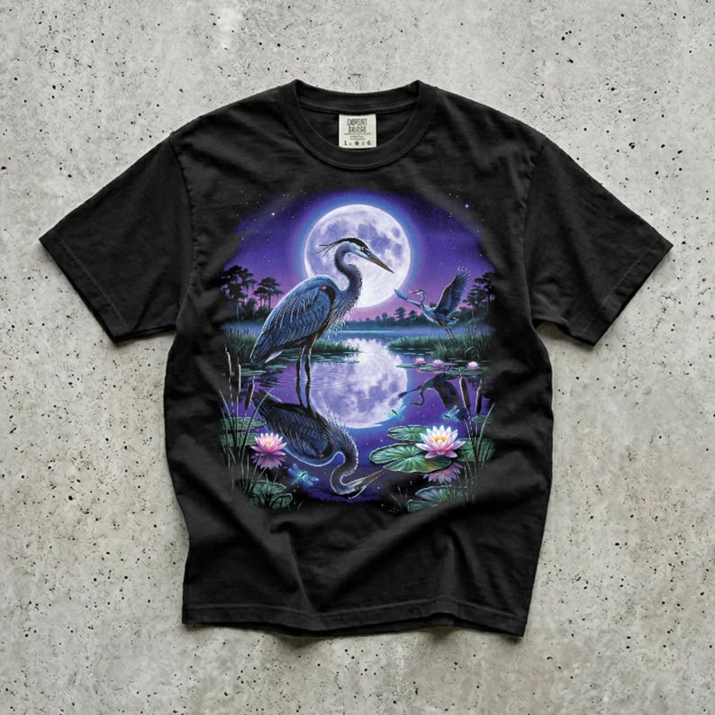 Black Comfort Colors garment-dyed t-shirt featuring a full-color 90s airbrush-style illustration of a great blue heron standing in a moonlit marsh with its reflection in violet water, a second heron in flight, pink water lilies, cattails, and a full moon in a star-scattered purple night sky — unisex graphic tee casually laid out on a concrete surface.