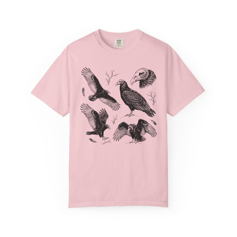 Light pink Comfort Colors t-shirt with a vintage engraved illustration of five vultures including one soaring with wings outstretched, one standing with wings half-spread, a close-up portrait showing bare head and hooked beak, one landing with wings fanned, and one spreading wings low in a dominance display, surrounded by bare winter branches and scattered feathers in an antique wildlife field guide etching style.