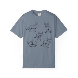 Blue jean graphic t-shirt featuring a vintage engraved style illustration of multiple fennec foxes arranged in different poses. The foxes are shown sitting, walking, and curled up, with detailed linework creating a naturalist wildlife illustration and desert animal aesthetic.