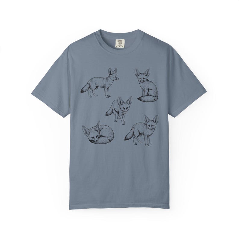 Blue jean graphic t-shirt featuring a vintage engraved style illustration of multiple fennec foxes arranged in different poses. The foxes are shown sitting, walking, and curled up, with detailed linework creating a naturalist wildlife illustration and desert animal aesthetic.