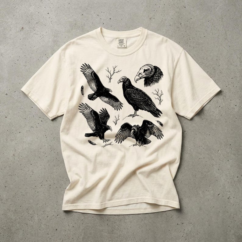 Ivory Comfort Colors t-shirt with a vintage engraved illustration of five vultures including one soaring with wings outstretched, one standing with wings half-spread, a close-up portrait showing bare head and hooked beak, one landing with wings fanned, and one spreading wings low in a dominance display, surrounded by bare winter branches and scattered feathers in an antique wildlife field guide etching style. Laid flat on a concrete surface.