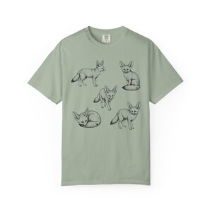 Bay green graphic t-shirt featuring a vintage engraved style illustration of multiple fennec foxes arranged in different poses. The foxes are shown sitting, walking, and curled up, with detailed linework creating a naturalist wildlife illustration and desert animal aesthetic.
