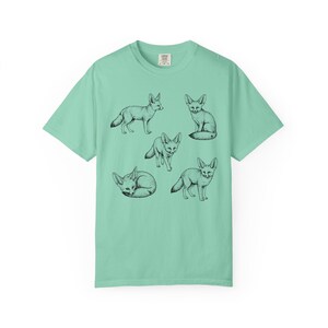 Island reef green graphic t-shirt featuring a vintage engraved style illustration of multiple fennec foxes arranged in different poses. The foxes are shown sitting, walking, and curled up, with detailed linework creating a naturalist wildlife illustration and desert animal aesthetic.