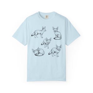 Light blue graphic t-shirt featuring a vintage engraved style illustration of multiple fennec foxes arranged in different poses. The foxes are shown sitting, walking, and curled up, with detailed linework creating a naturalist wildlife illustration and desert animal aesthetic.
