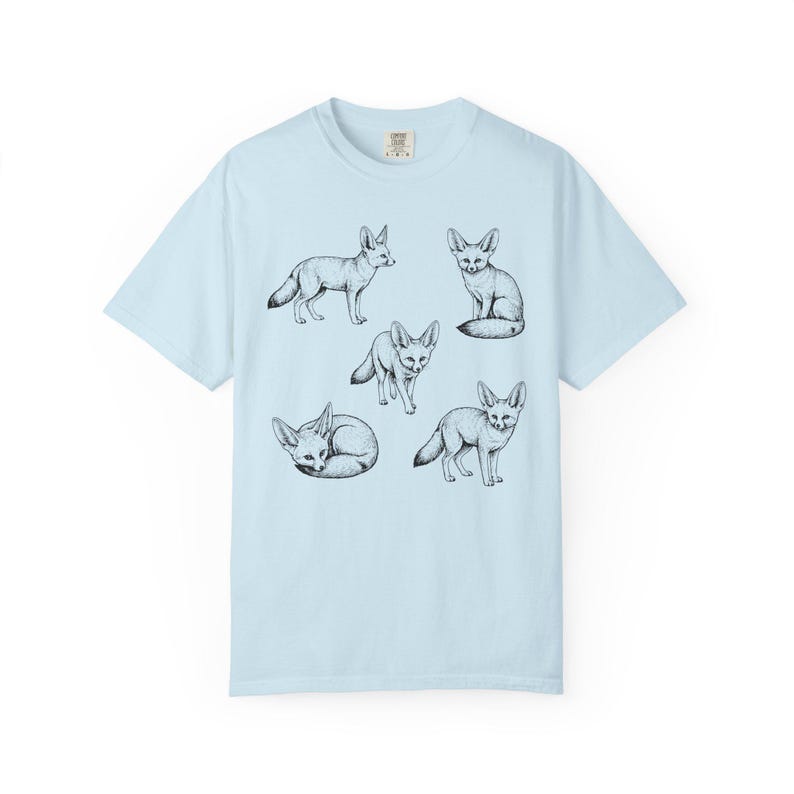 Light blue graphic t-shirt featuring a vintage engraved style illustration of multiple fennec foxes arranged in different poses. The foxes are shown sitting, walking, and curled up, with detailed linework creating a naturalist wildlife illustration and desert animal aesthetic.