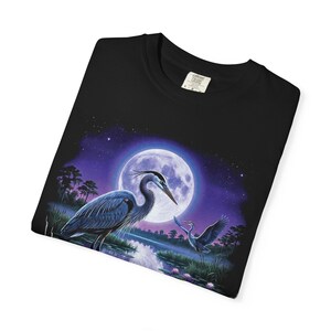 Black Comfort Colors garment-dyed t-shirt featuring a full-color 90s airbrush-style illustration of a great blue heron standing in a moonlit marsh with its reflection in violet water, a second heron in flight, pink water lilies, cattails, and a full moon in a star-scattered purple night sky — unisex graphic tee folded into a neat square.