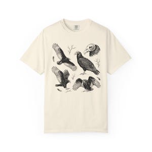 Ivory Comfort Colors t-shirt with a vintage engraved illustration of five vultures including one soaring with wings outstretched, one standing with wings half-spread, a close-up portrait showing bare head and hooked beak, one landing with wings fanned, and one spreading wings low in a dominance display, surrounded by bare winter branches and scattered feathers in an antique wildlife field guide etching style.