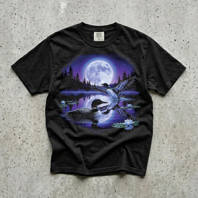 Black Comfort Colors garment-dyed t-shirt featuring a full-color 90s airbrush-style circular vignette illustration of a common loon swimming on a moonlit violet northern lake with a second loon in flight, blue water lilies, pine silhouettes, and a full moon — unisex graphic tee casually laid out on a concrete surface.