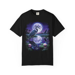 Black Comfort Colors garment-dyed t-shirt featuring a full-color 90s airbrush-style illustration of a great blue heron standing in a moonlit marsh with its reflection in violet water, a second heron in flight, pink water lilies, cattails, and a full moon in a star-scattered purple night sky — unisex graphic tee.