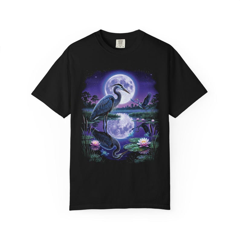 Black Comfort Colors garment-dyed t-shirt featuring a full-color 90s airbrush-style illustration of a great blue heron standing in a moonlit marsh with its reflection in violet water, a second heron in flight, pink water lilies, cattails, and a full moon in a star-scattered purple night sky — unisex graphic tee.