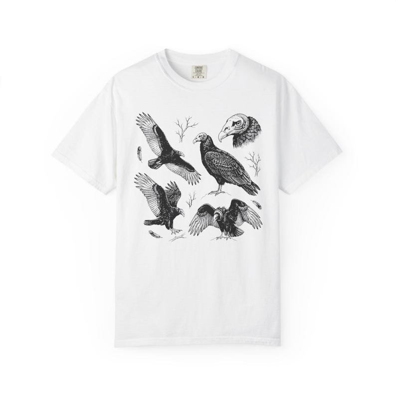 White Comfort Colors t-shirt with a vintage engraved illustration of five vultures including one soaring with wings outstretched, one standing with wings half-spread, a close-up portrait showing bare head and hooked beak, one landing with wings fanned, and one spreading wings low in a dominance display, surrounded by bare winter branches and scattered feathers in an antique wildlife field guide etching style.