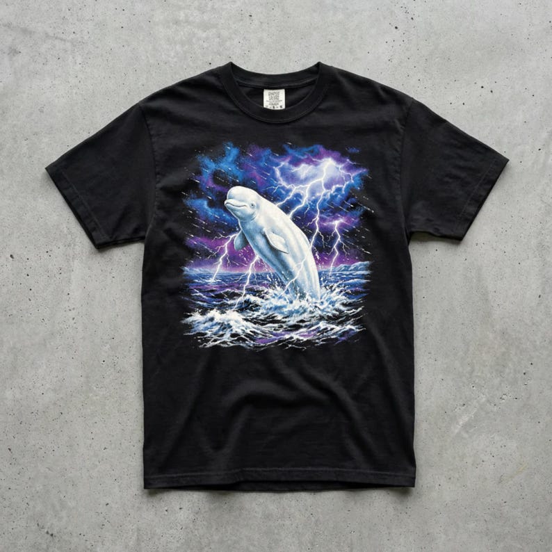 Black Comfort Colors garment-dyed t-shirt featuring a full-color 90s airbrush-style illustration of a white beluga whale breaching from a dark storm ocean beneath forking white lightning bolts and deep cobalt and purple storm clouds, laid flat on a concrete surface — unisex graphic tee.