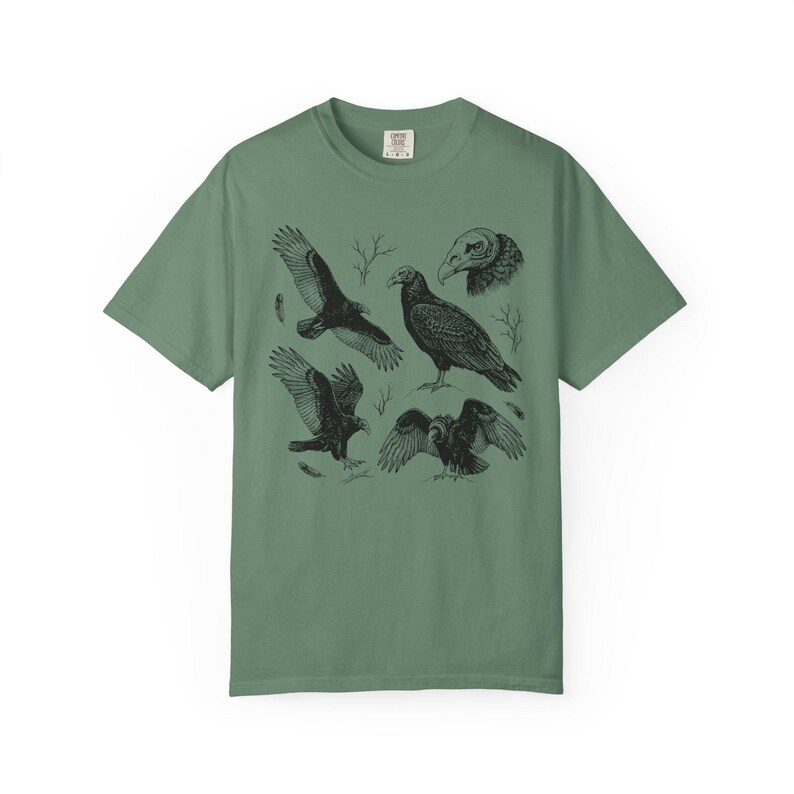 Light green Comfort Colors t-shirt with a vintage engraved illustration of five vultures including one soaring with wings outstretched, one standing with wings half-spread, a close-up portrait showing bare head and hooked beak, one landing with wings fanned, and one spreading wings low in a dominance display, surrounded by bare winter branches and scattered feathers in an antique wildlife field guide etching style.